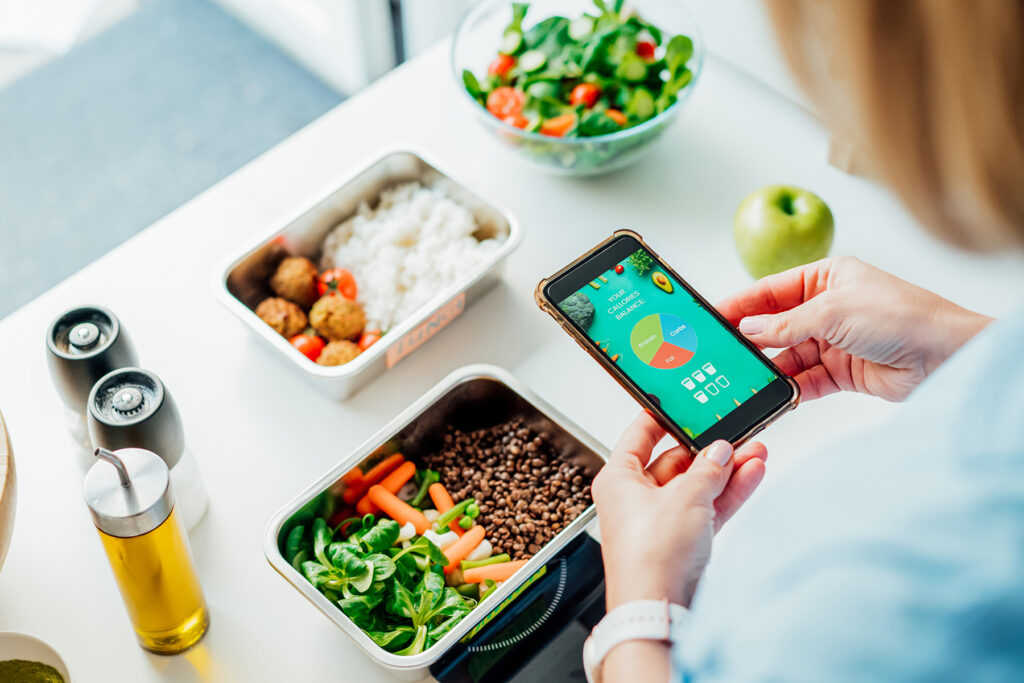 Healthy diet plan for weight loss, daily ready meal menu. Woman using meal tracker app on phone while weighing lunch box cooked in advance on kitchen scale. Balanced portion with dish. Pre-cooking