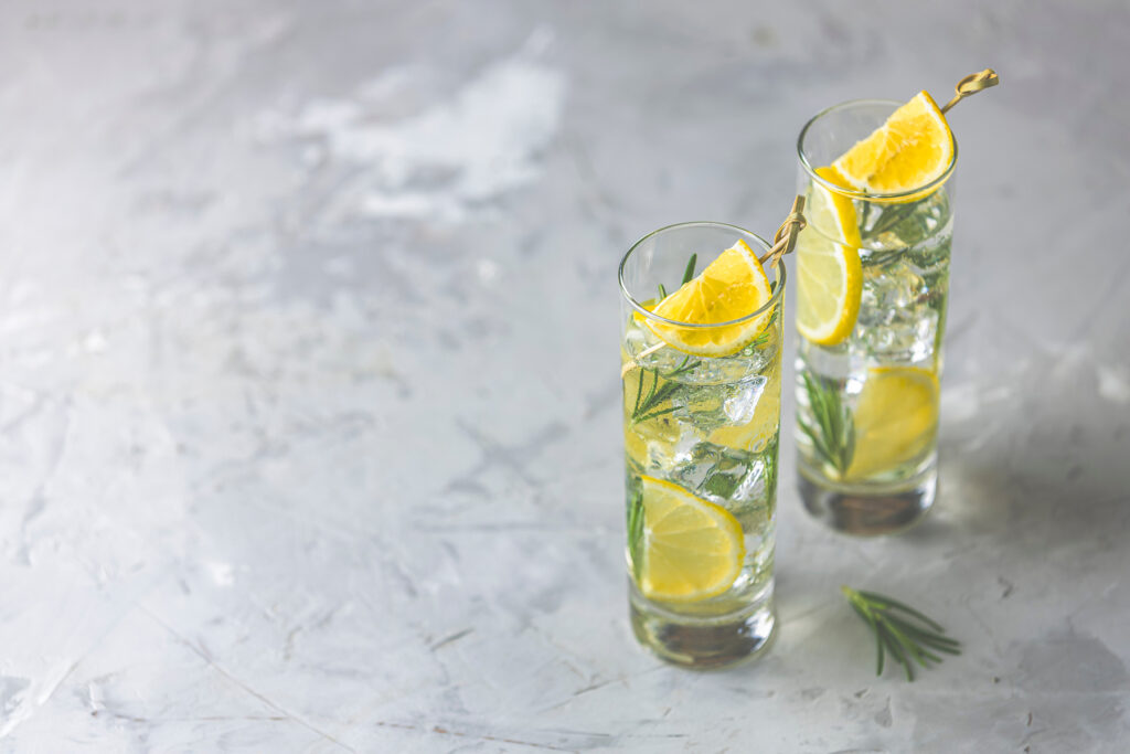Two glasses of refreshing lemon lime drink with ice cubes in glass goblets with water drops against a light gray background. Summer fresh lemon soda cocktail with rosemary, selective focus