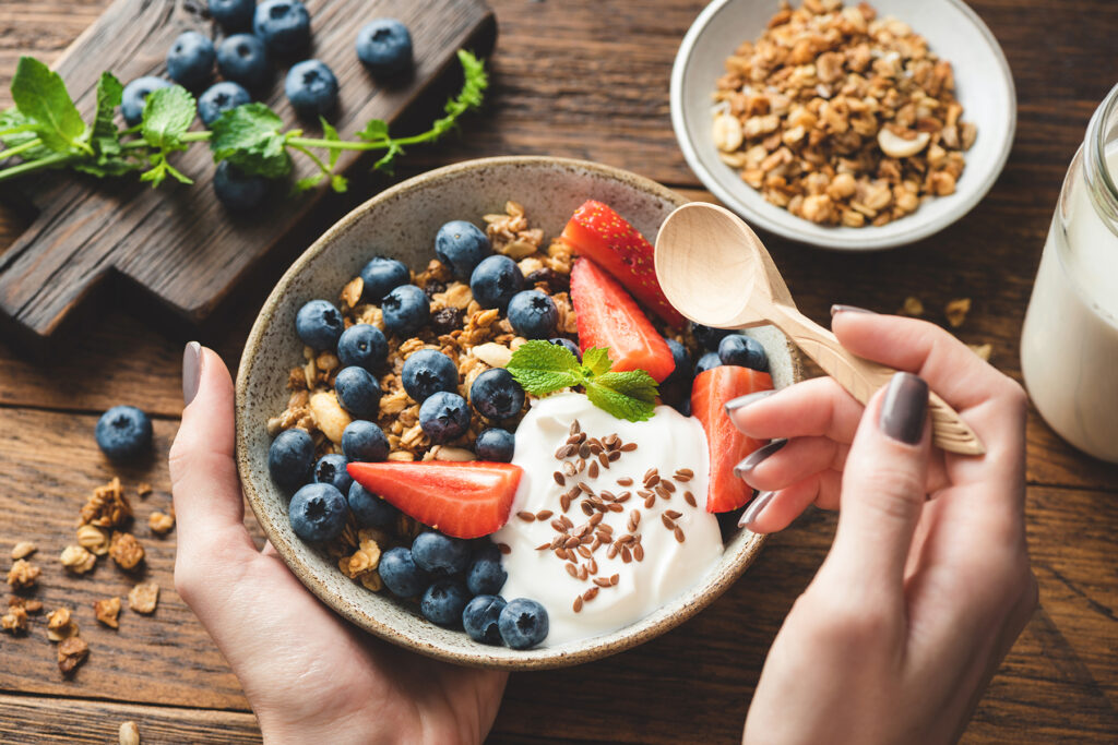 Eating granola with greek yogurt and berries. Female hands holding bowl of healthy breakfast cereals granola with fruits and yogurt. Concept of clean eating, dieting